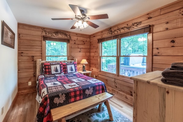A cozy wooden cabin bedroom with a bed, a ceiling fan, and large windows.