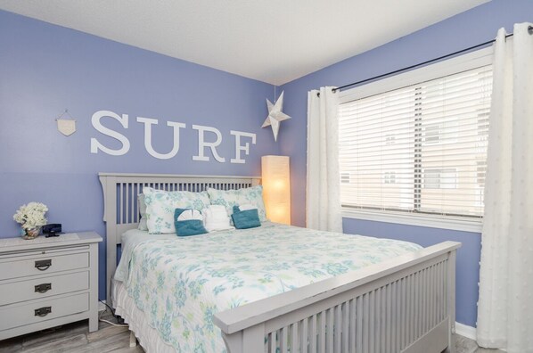 A bedroom with a bed, a nightstand, a wall-mounted lamp, and a window with blinds. - First Floor with Ocean Views from Every Room (Carolina Beach)