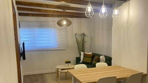 A modern living room with a wooden dining table, white chairs, a green sofa with cushions, and a large window with blinds.