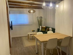 Uma sala de estar moderna com uma mesa de jantar de madeira, cadeiras brancas, um sofá verde com almofadas e uma janela grande com persianas.