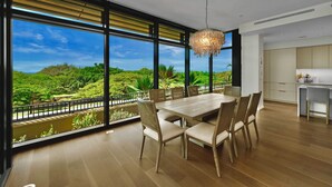 Un espace repas comprenant une grande table et des chaises, un luminaire suspendu et une vue sur une végétation luxuriante grâce à de grandes fenêtres.