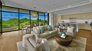 Un vaste salon doté d’une grande fenêtre donnant sur un paysage luxuriant, une cuisine moderne avec des meubles blancs et un coin repas comprenant une table et des chaises hautes.