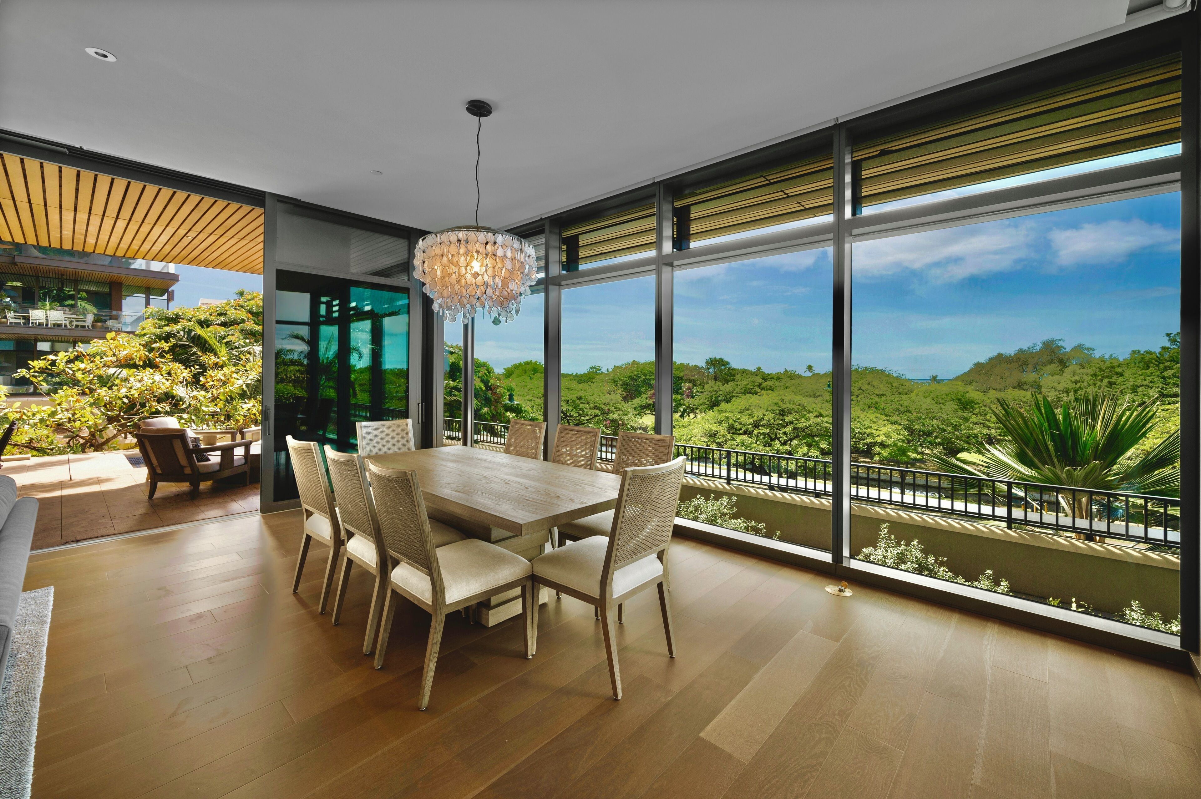 Un comedor amplio con una mesa grande y sillas, ventanas panorámicas que ofrecen vistas a un follaje exuberante, y una lámpara moderna.