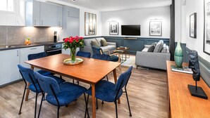 A modern kitchen with grey cabinets, a dining area with a wooden table and blue chairs, and a living room with a grey sofa and a flat-screen TV. - LivinParis | Luxurious 2 Bedroom Opera VI (Paris)