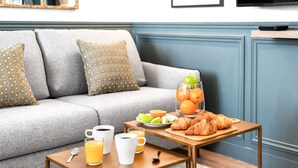 A modern living room with a grey sofa, patterned cushions, a wooden coffee table with breakfast items, and a blue wall with a television. - LivinParis | Luxurious 2 Bedroom Opera VI (Paris)