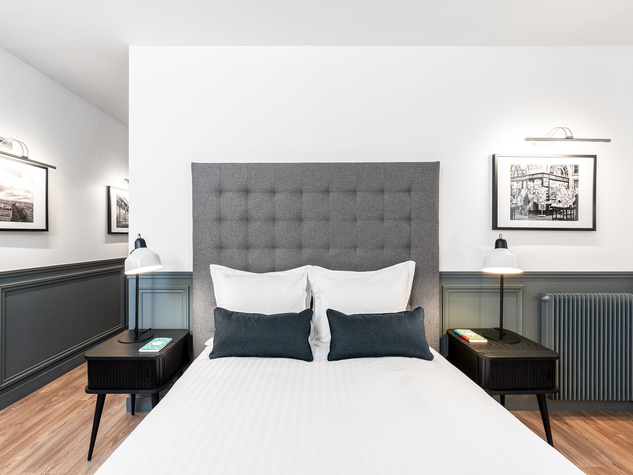 A modern hotel room with a large bed, two bedside tables, a radiator, and framed pictures on the walls.
