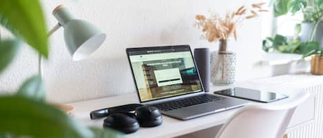 A laptop on a desk with a website open, a lamp, headphones, and a vase with dried flowers.
