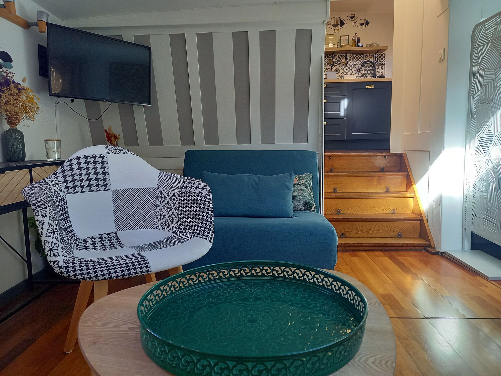 A living room with a blue sofa, a patterned armchair, a green decorative bowl, a flat-screen TV, and a staircase leading to a kitchen.