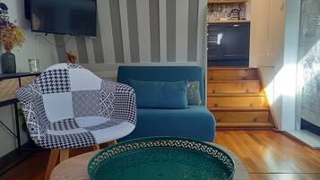A living room with a blue sofa, a patterned armchair, a green decorative bowl, a flat-screen TV, and a staircase leading to a kitchen.