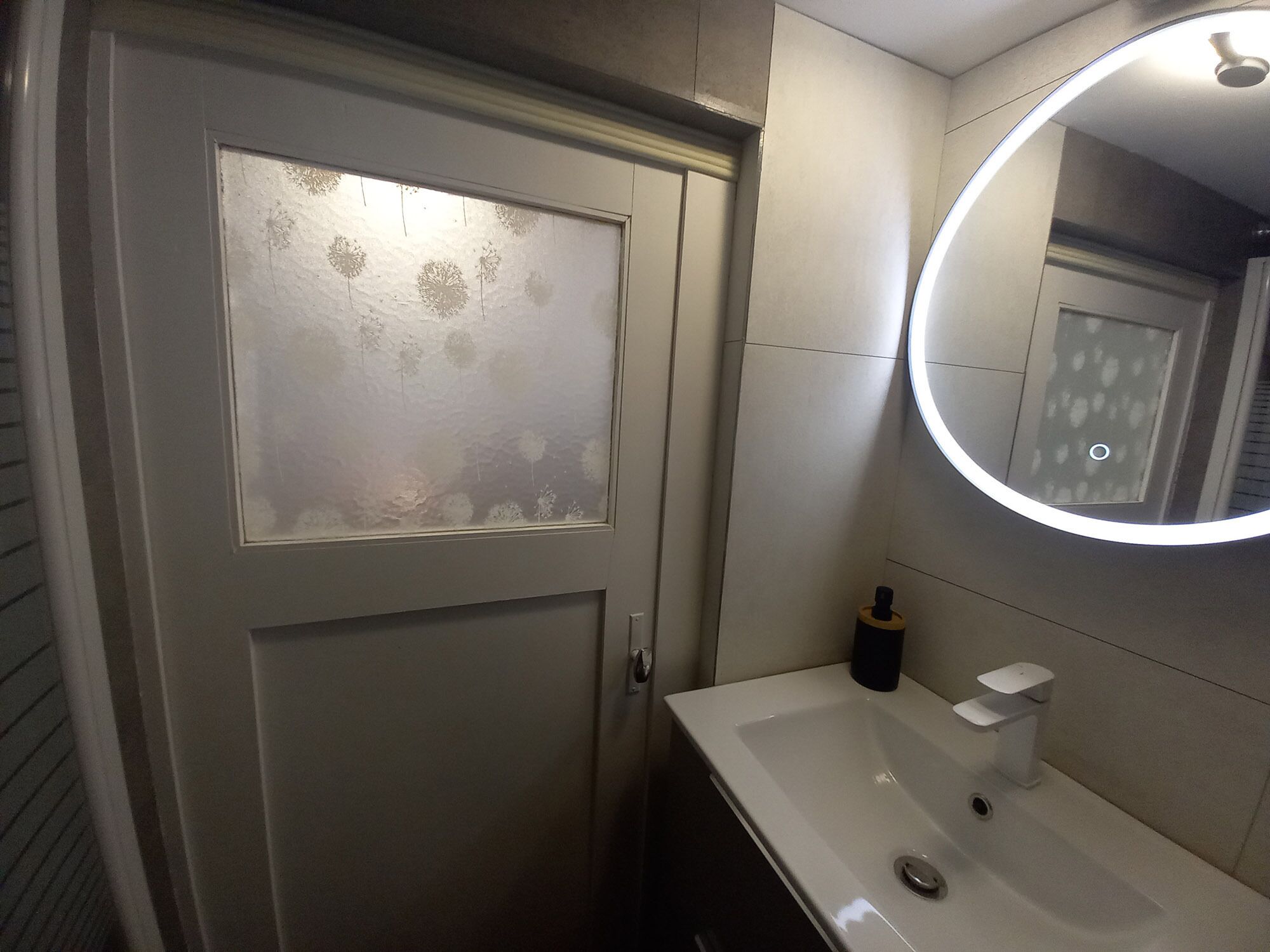 A bathroom with a frosted glass door, a white sink with a mirror, and a round mirror with a light.