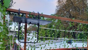 Une terrasse en bois avec une pergola, un pot de fleurs et, au loin, la vue d’un bâtiment.