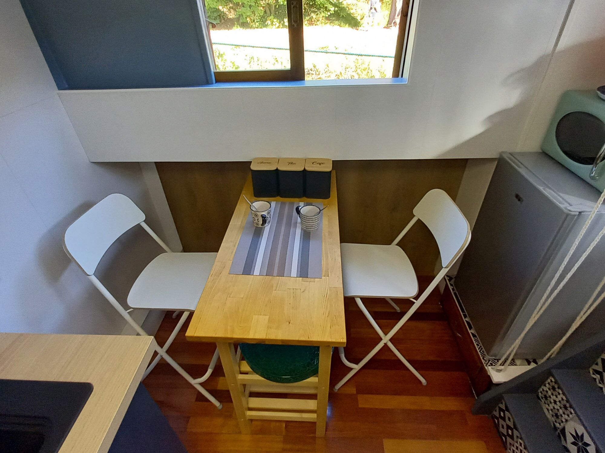 Een kleine kamer met een houten tafel, twee witte stoelen, een klein raam en een radiator.