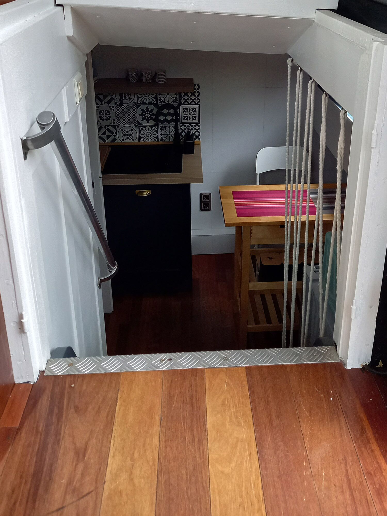 Una habitación con suelo de madera, un escritorio con mantel de rayas rosa y rojo, una silla y una cama con cabecera blanca. Se observa una pared con diseño detrás del escritorio y una reja metálica frente a la cama.