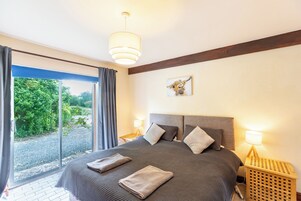 Quarto com cama grande, vista para um jardim através de uma porta deslizante e um vigamento de madeira no teto.
