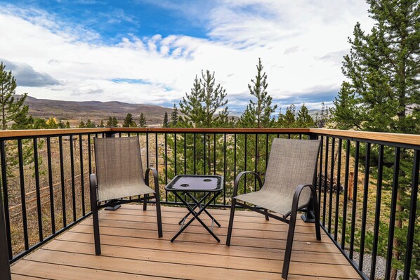 A wooden deck with two chairs and a small table, surrounded by a metal railing, overlooking a mountainous landscape.