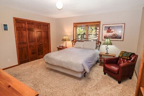 A bedroom with a large bed, a wooden wardrobe, a nightstand with a lamp, a framed picture on the wall, and a red leather chair.