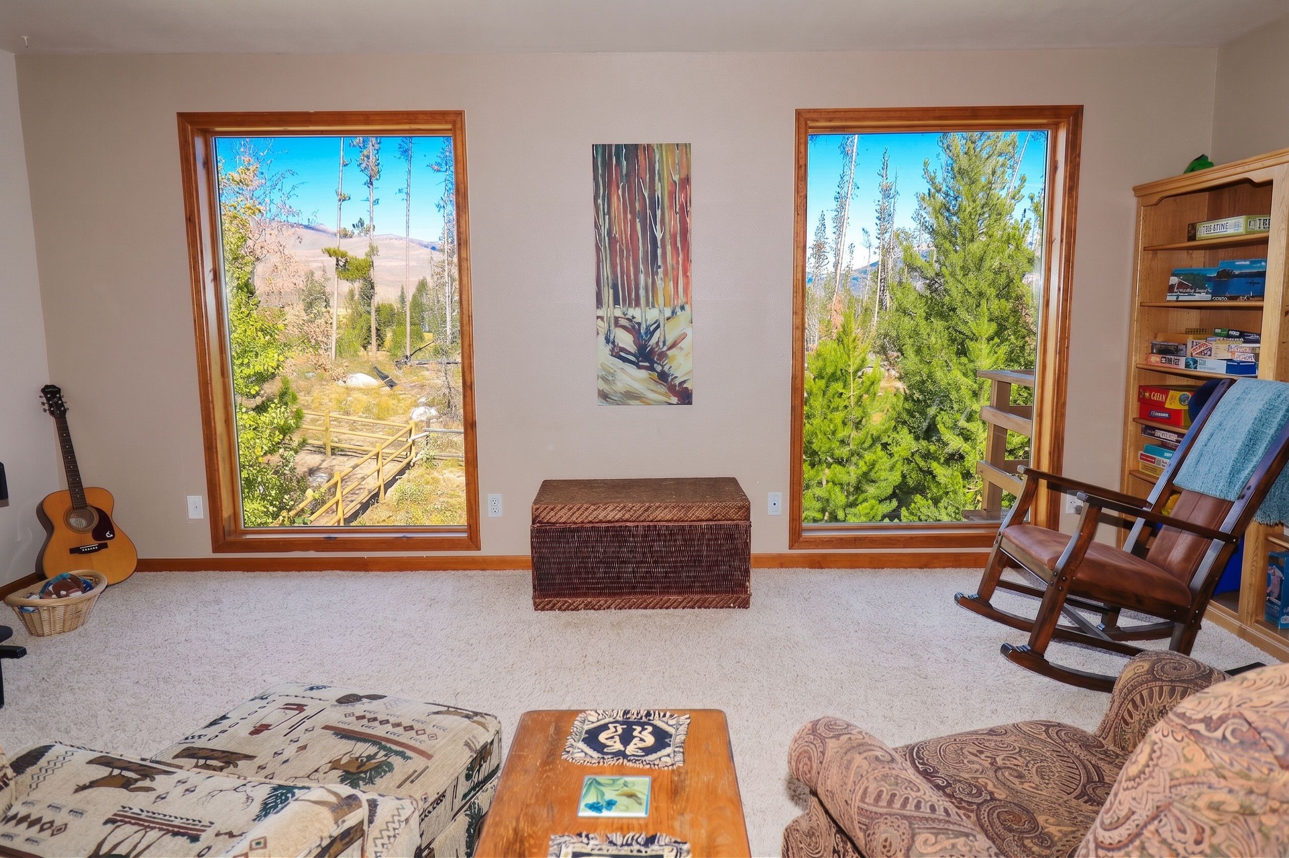 A living room with a view of a forest through large windows, a guitar, a rocking chair, and a bookshelf.