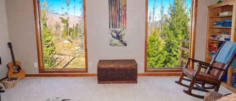 A living room with a view of a forest through large windows, a guitar, a rocking chair, and a bookshelf.