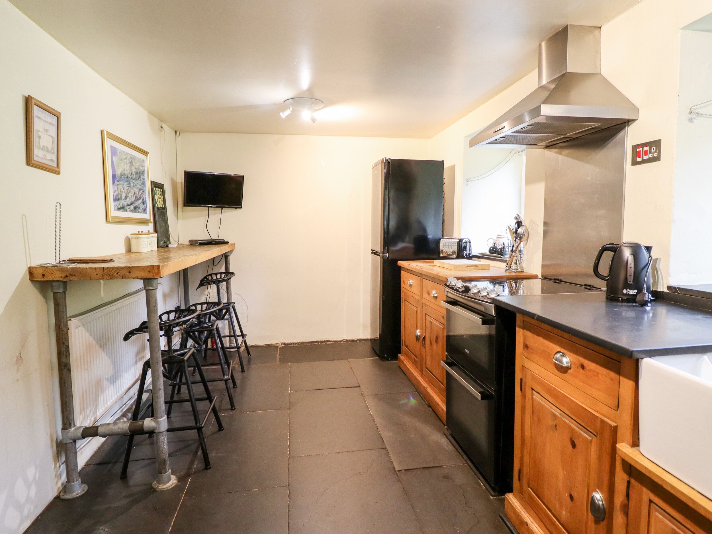 A compact kitchen with a refrigerator, stove, and sink. There is a wooden bar counter with stools and a television mounted on the wall.