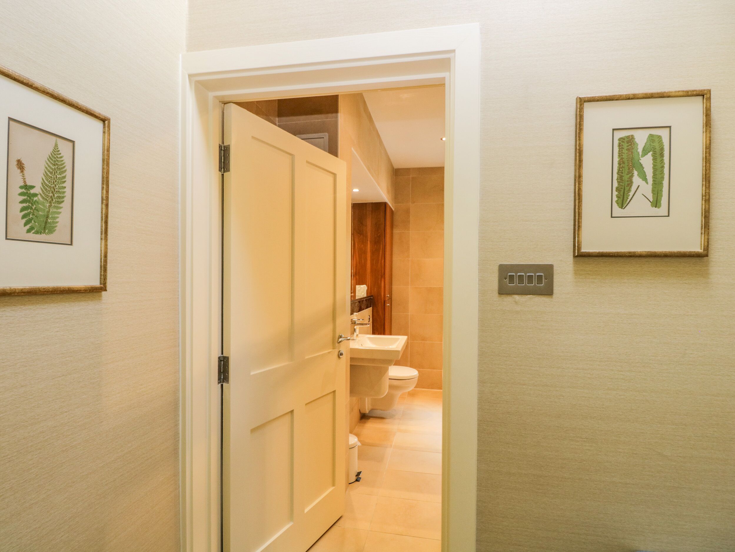 A hallway leading to a bathroom with a toilet and sink. There are framed botanical prints on the walls.