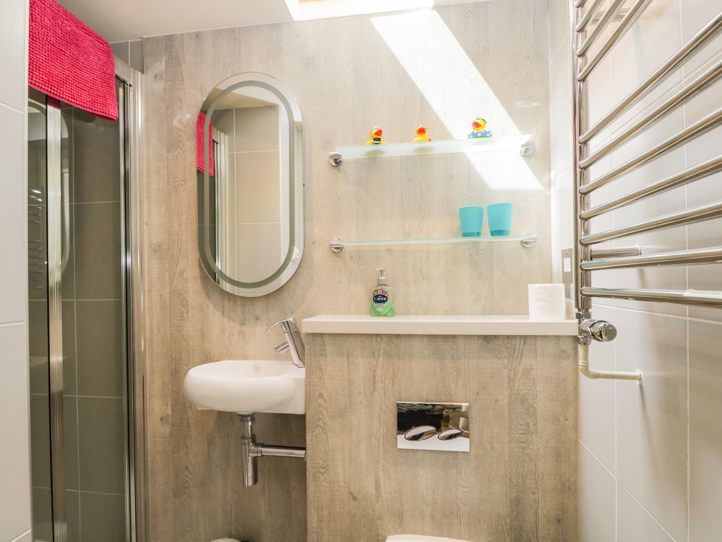 Une salle de bain avec une douche à parois de verre, un lavabo blanc et un miroir. On y trouve des étagères avec des articles de toilette et un porte-serviettes.