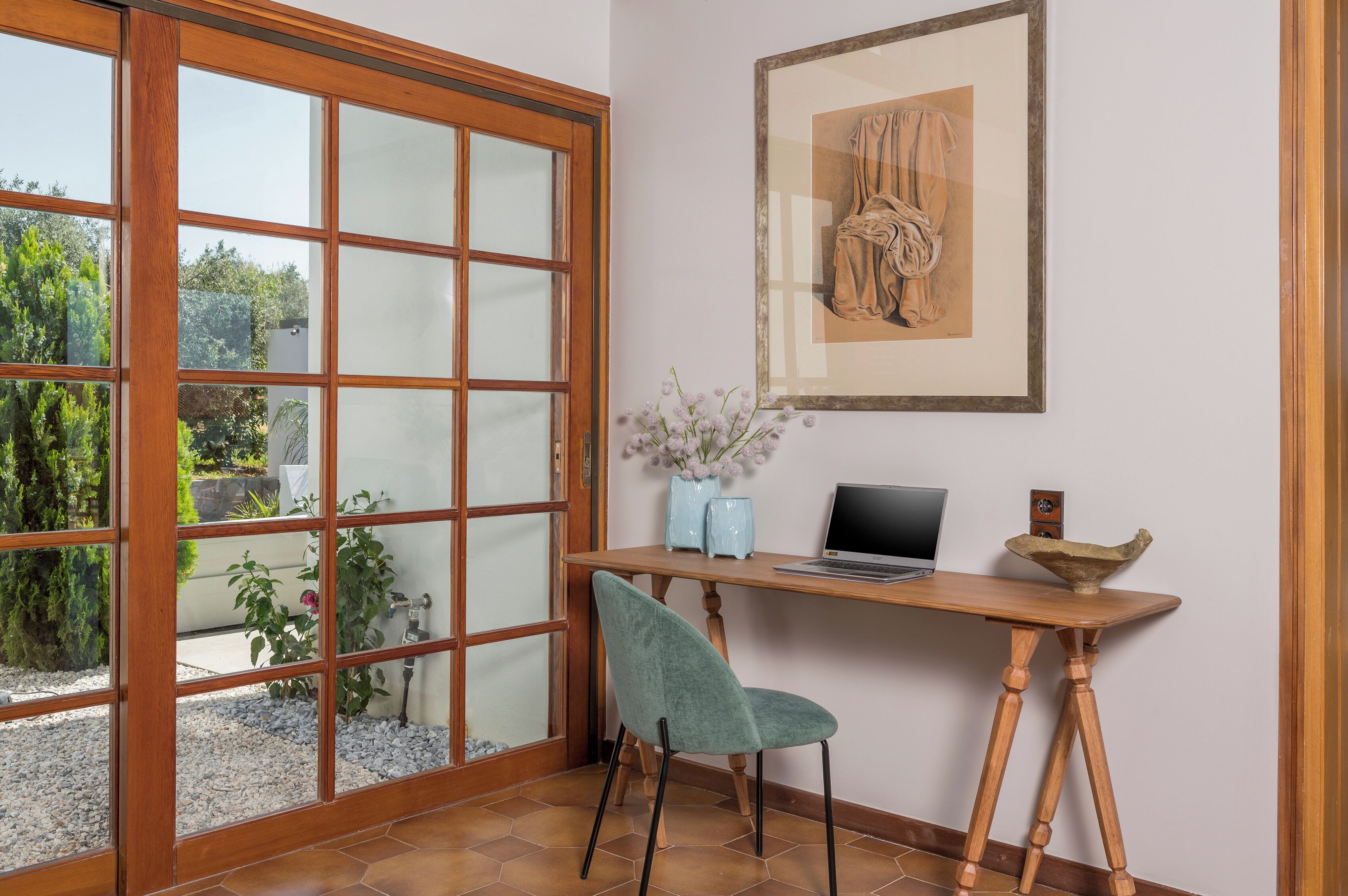 Et værelse med et træskrivebord, en bærbar computer, en vase med blomster og et indrammet kunstværk på væggen.
