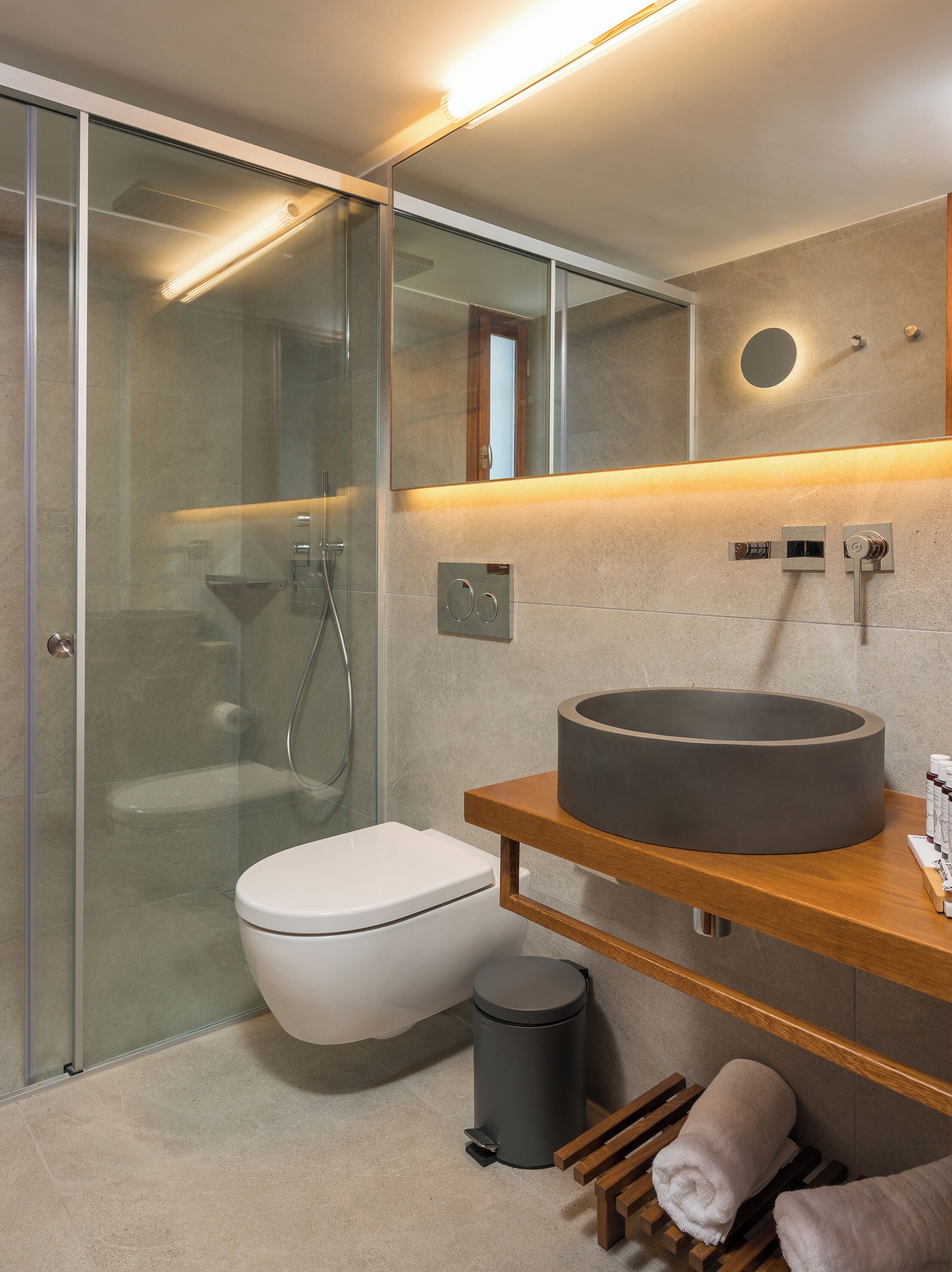 Un baño moderno con mampara de cristal, inodoro blanco, un lavabo grande y redondo, y un mueble de tocador de madera.