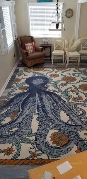 A room with a large octopus-patterned rug, wicker chairs, a small side table, and a hanging light fixture.