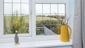 A window with a view of a green landscape, a yellow vase with branches, and a small figurine on the windowsill. - Pond Farm View (Saltburn-by-the-Sea)