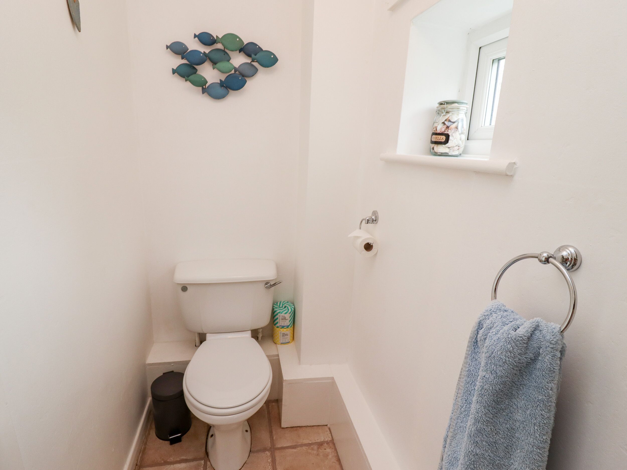 A small bathroom with a toilet, a trash bin, a towel rack, and a shelf with a jar.