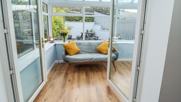 A conservatory with a wooden floor, a grey sofa with yellow cushions, and a view of a garden through large glass doors. - Pond Farm View (Saltburn-by-the-Sea)