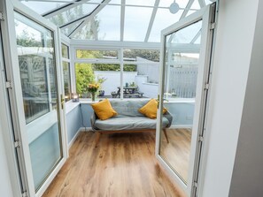 Ein Wintergarten mit Holzboden, einem grauen Sofa mit gelben Kissen und Blick auf einen Garten durch große Glasüren.