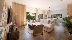 Amplia sala de estar con sofá, mesa de centro y una silla. Se ve una ventana grande con cortinas, un televisor y un área de cocina al otro lado.