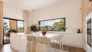 Una cocina moderna con una isla grande, taburetes blancos de bar, un frutero y vistas al exterior.