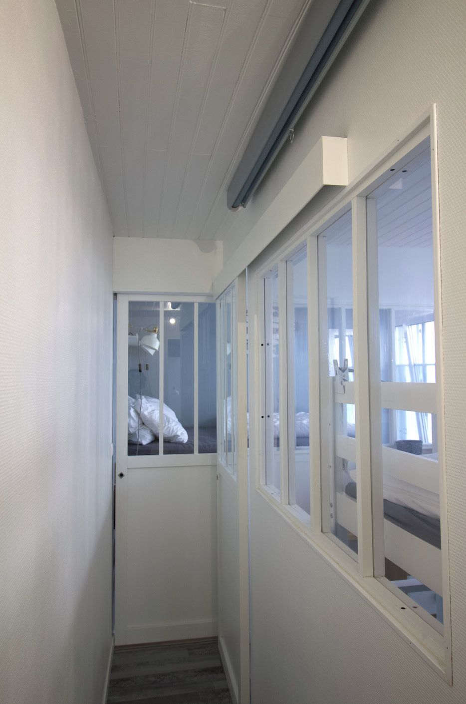 Ein schmaler Flur mit einer Tür zu einem Zimmer mit Bett und Fenster.