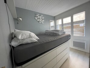 Une chambre à coucher avec un lit, des oreillers, une lampe fixée au mur et une fenêtre donnant sur l’extérieur.