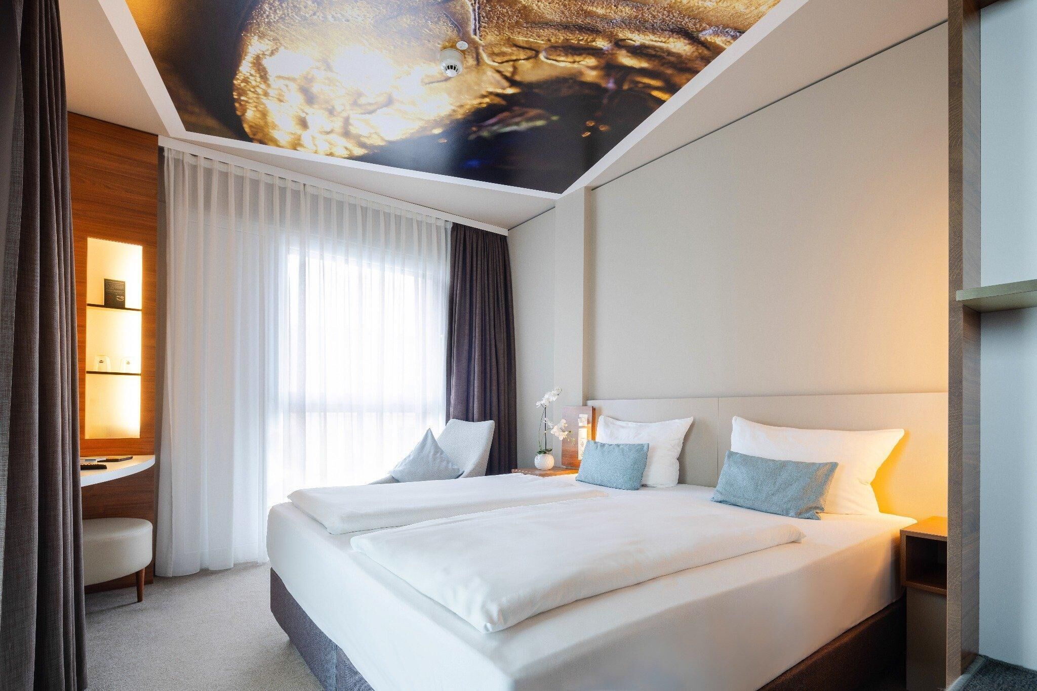 A hotel room with two beds, a desk, a ceiling mural, and a large window with curtains.