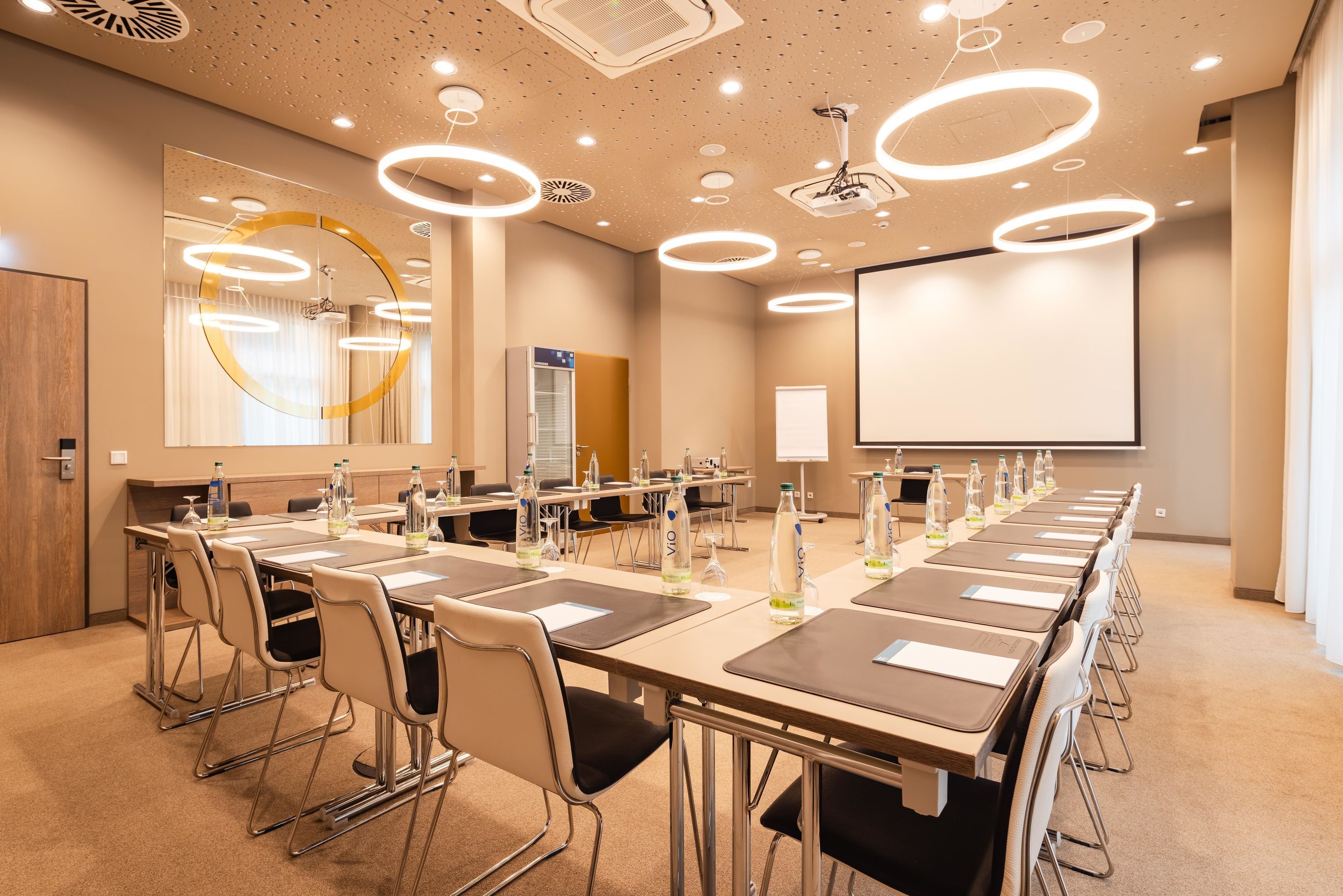 A conference room with a long table, chairs, a projector screen, and a large mirror.