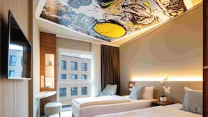 Habitación de hotel con dos camas, un escritorio, un televisor y un ventanal con vistas a un edificio.
