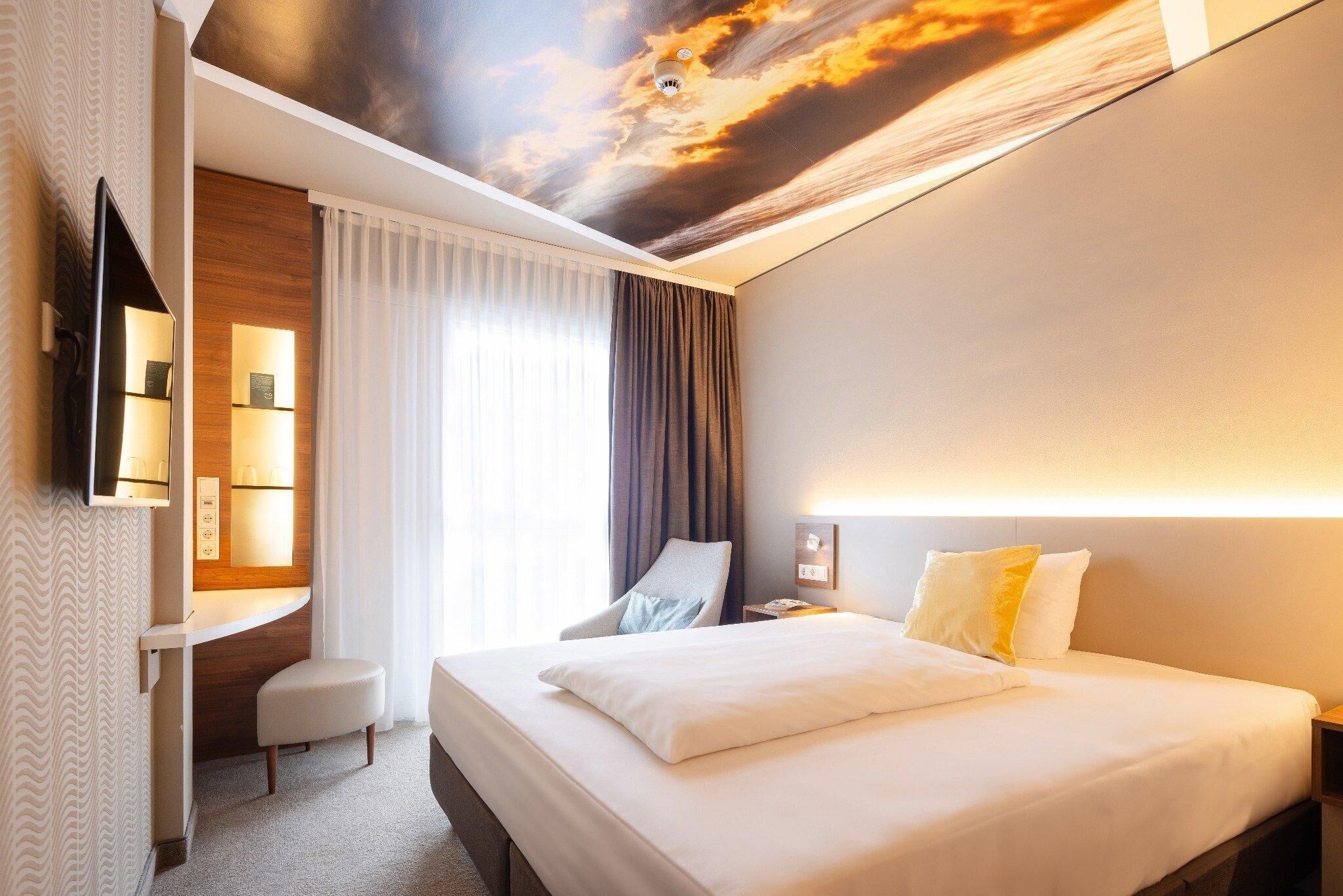 Ein Hotelzimmer mit Bett, Schreibtisch, Stuhl, Fernseher und Wandgemälde an der Decke.