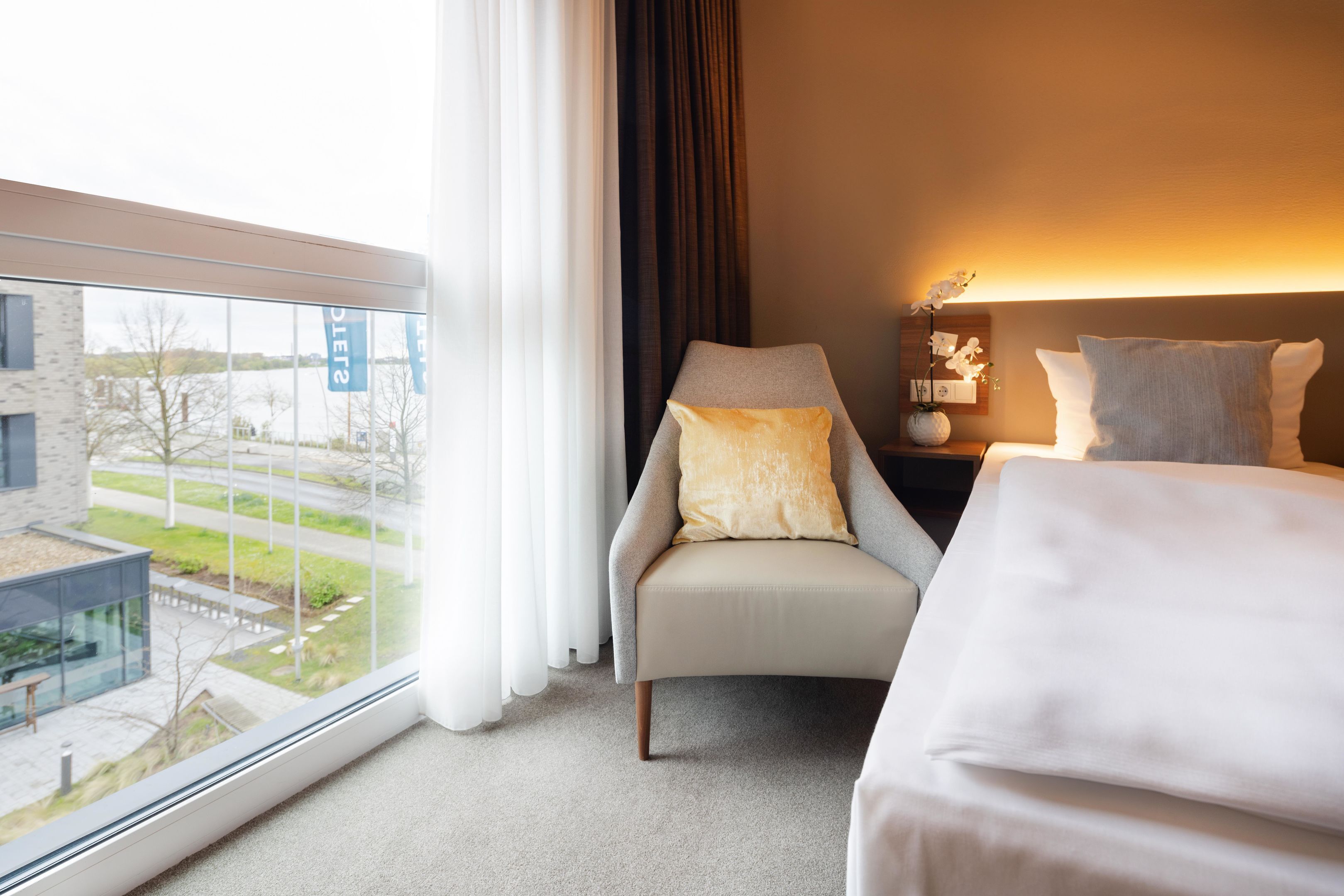 A hotel room with a view of a building outside, a chair with a pillow, a bedside table with a lamp, and a bed.