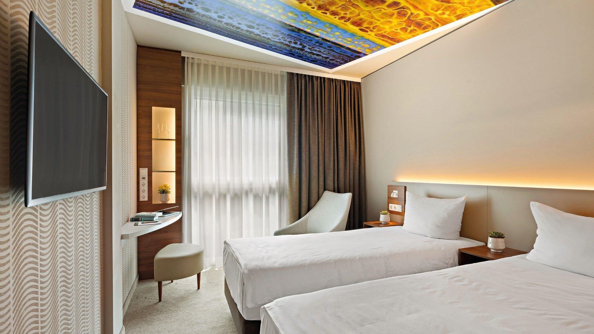 A hotel room with two beds, a desk, a chair, a TV, and a ceiling mural.