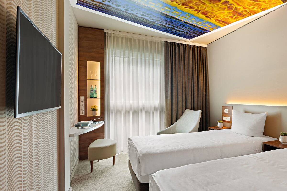 A hotel room with two beds, a desk, a chair, a TV, and a ceiling with a unique light design.