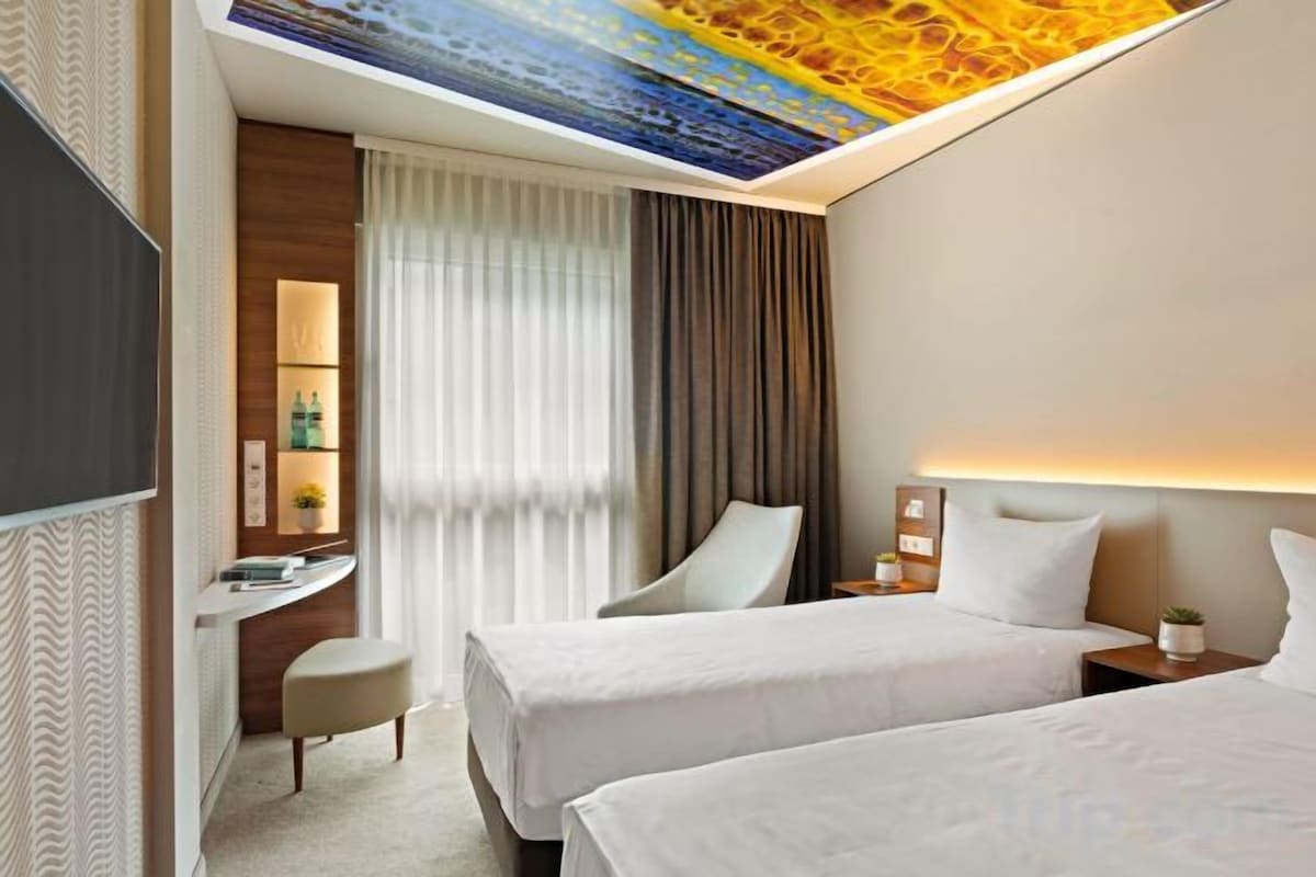 A hotel room with two beds, a desk, a chair, and a ceiling light fixture.