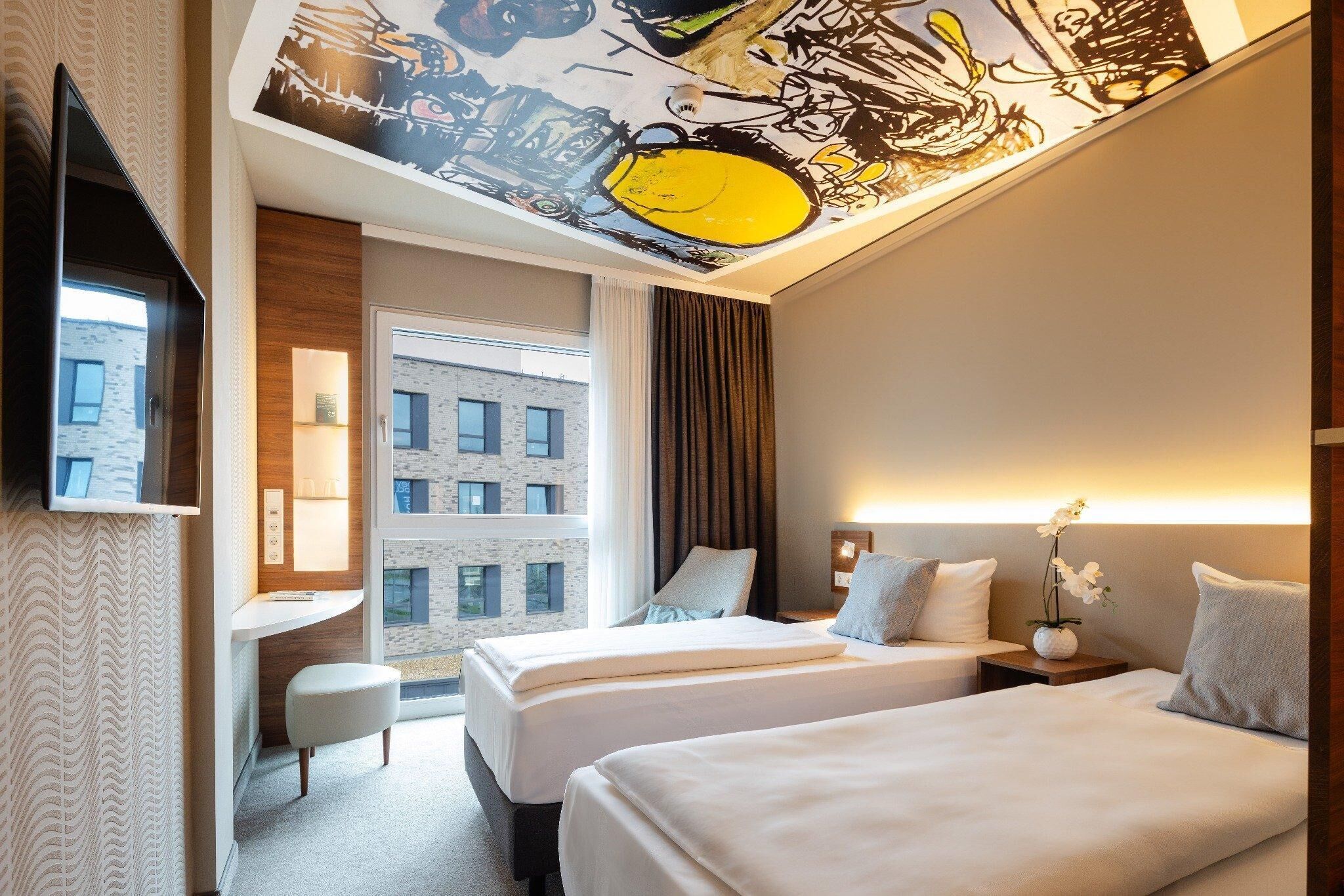 A hotel room with two beds, a desk, a chair, a TV, and a large window with a view of a building.
