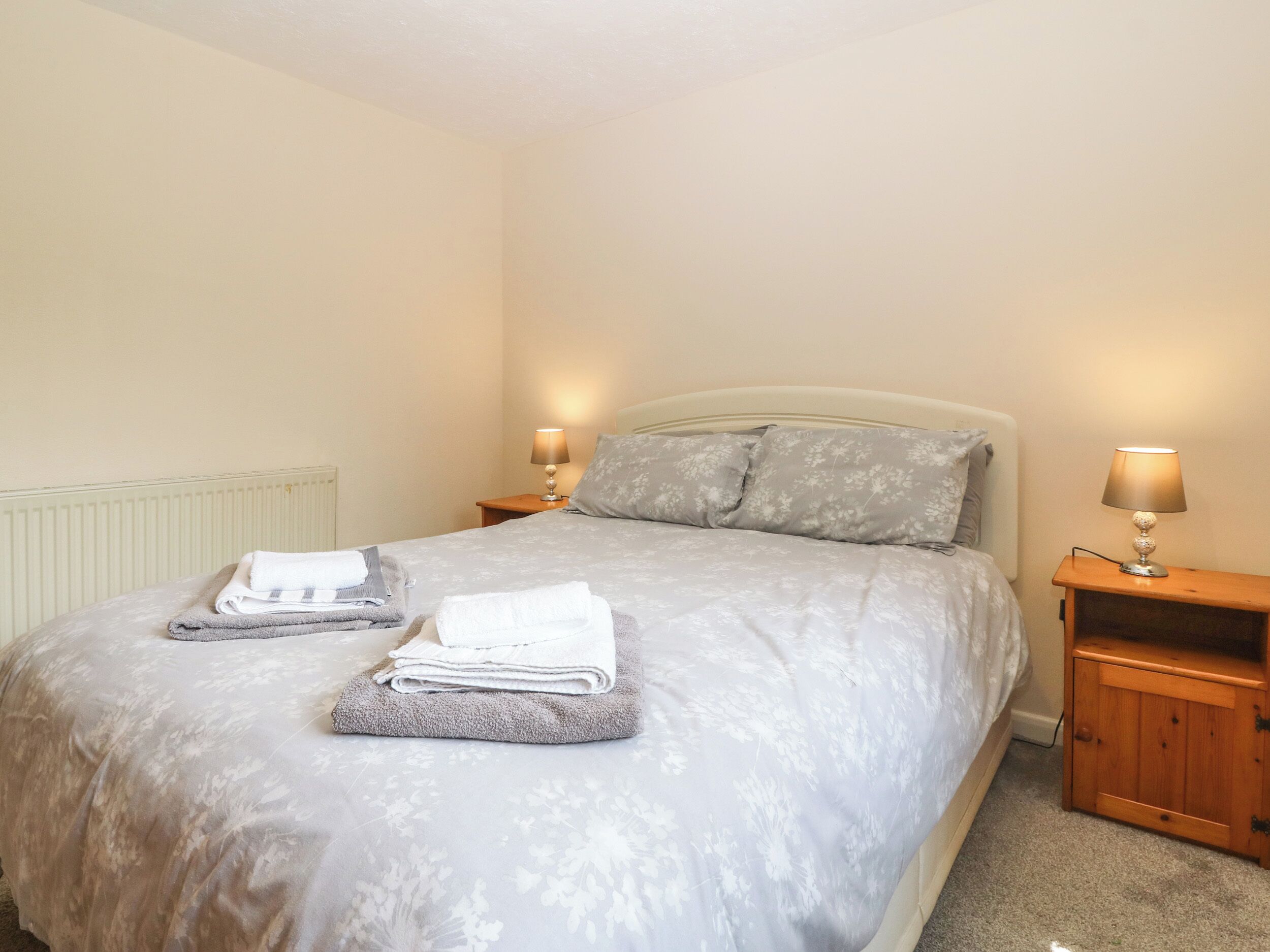 A neatly made bed with a floral-patterned bedspread, two pillows, and folded towels on the side. There are two bedside lamps on wooden nightstands.
