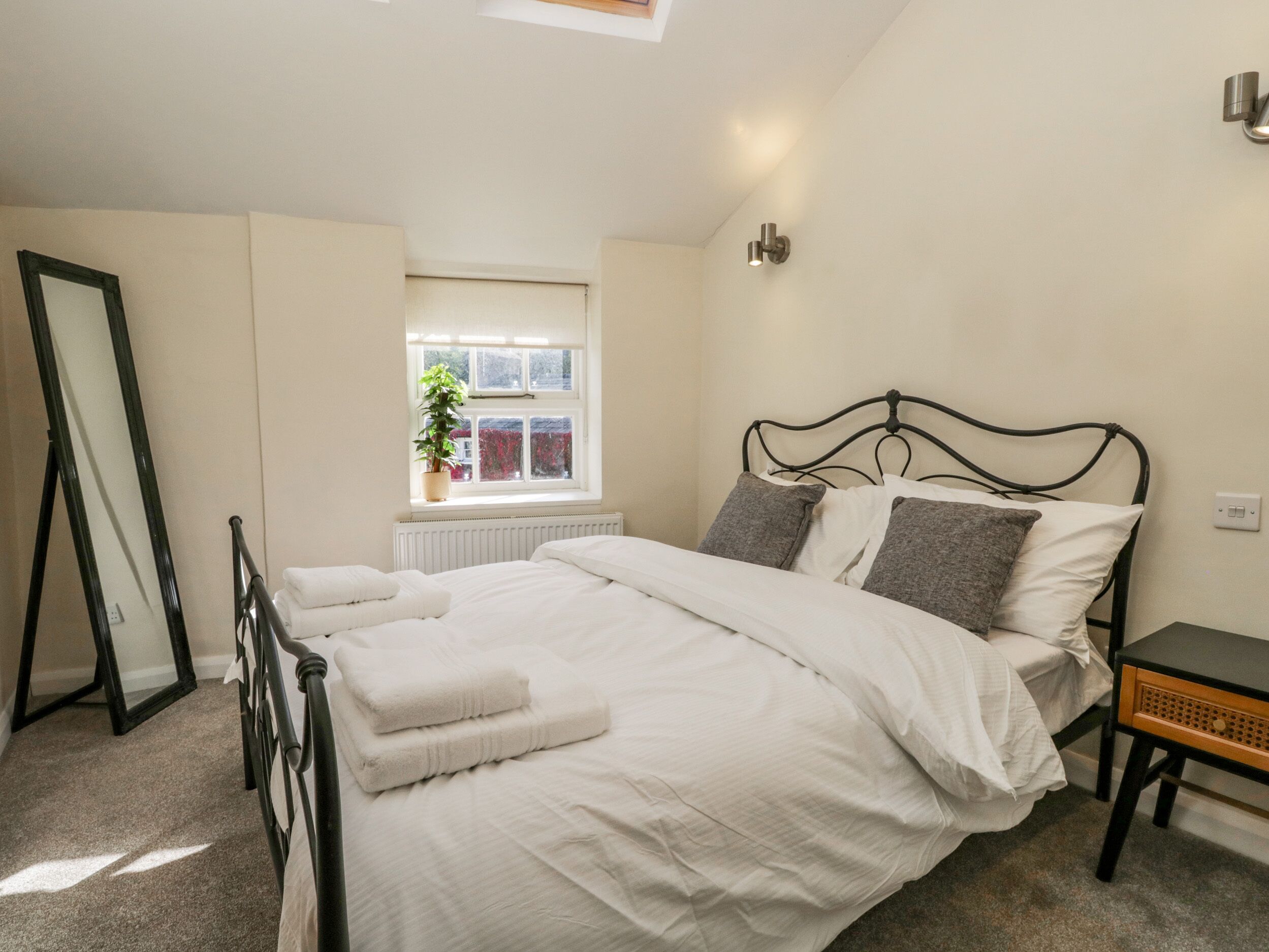 A neatly made bed with white linens and two pillows, a bedside table with a lamp, a window with a view of red flowers, and a radiator under the window.