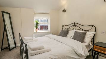 A neatly made bed with white linens and two pillows, a bedside table with a lamp, a window with a view of red flowers, and a radiator under the window.