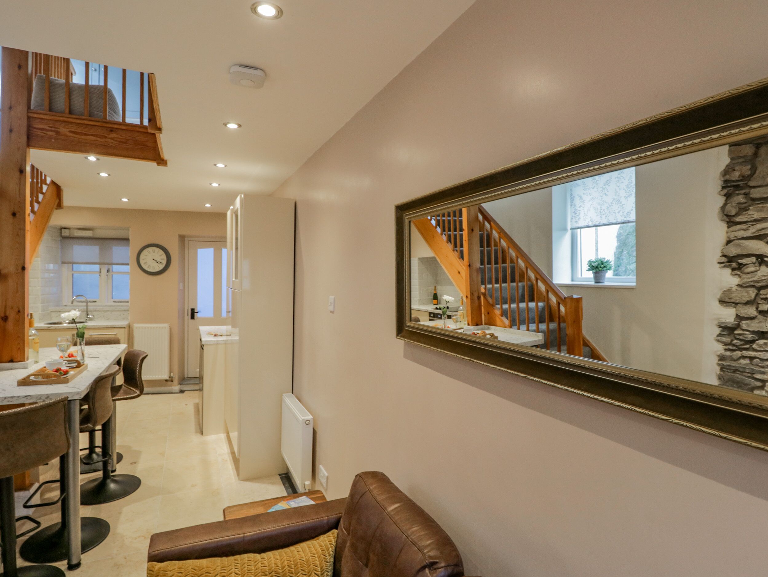 A modern kitchen with a wooden staircase, a dining area with bar stools, and a large mirror reflecting the staircase.