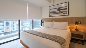 A modern hotel room with a large bed, a view of the city through floor-to-ceiling windows, a framed artwork above the bed, and a nightstand with a lamp.